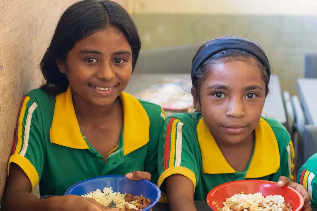 Two Timorese girls smile while wearing green school uniforms and holding bowls of food.